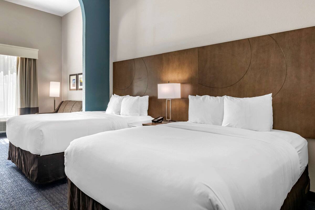 Two queen beds in a hotel room with a wooden headboard, a bedside table with a lamp, and a large window with curtains.