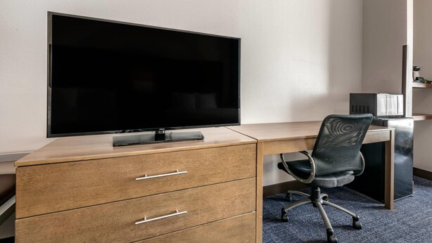 A hotel room with a wooden desk, a large flat-screen TV, a black office chair, and a microwave.