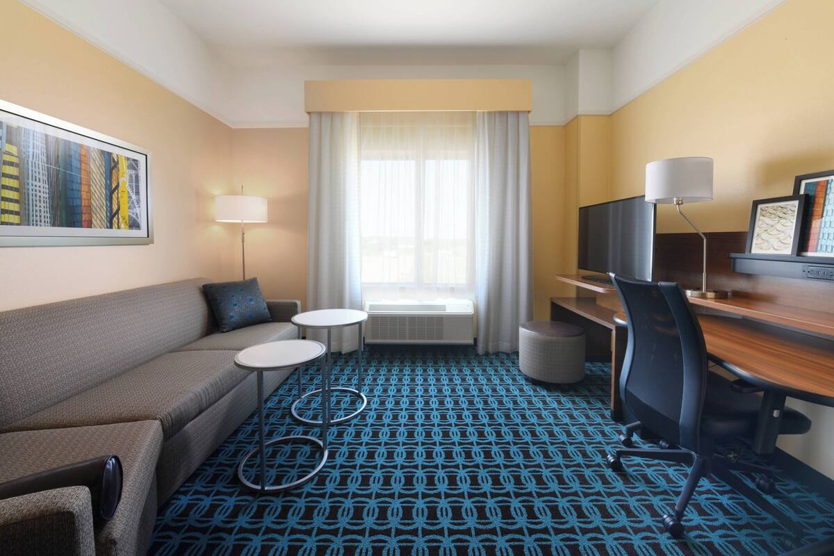 Een hotelkamer met een bank, twee kleine ronde tafels, een bureau met stoel, een televisie en een groot raam met gordijnen.