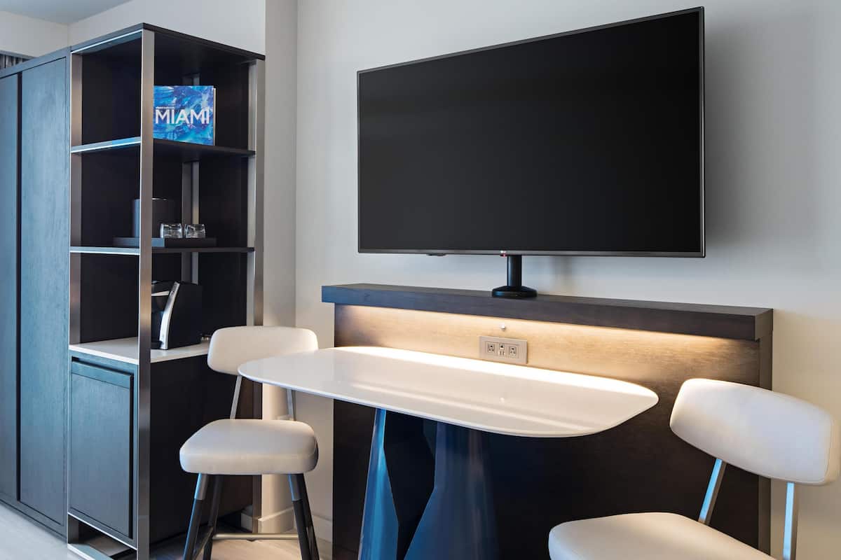 A modern hotel room with a flat-screen TV mounted on the wall, a built-in bench with stools, and a minibar with a 'MIAMI' sign.