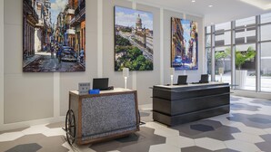 A hotel reception area with a check-in counter, a desk with a laptop, and three large paintings of cityscapes on the wall.