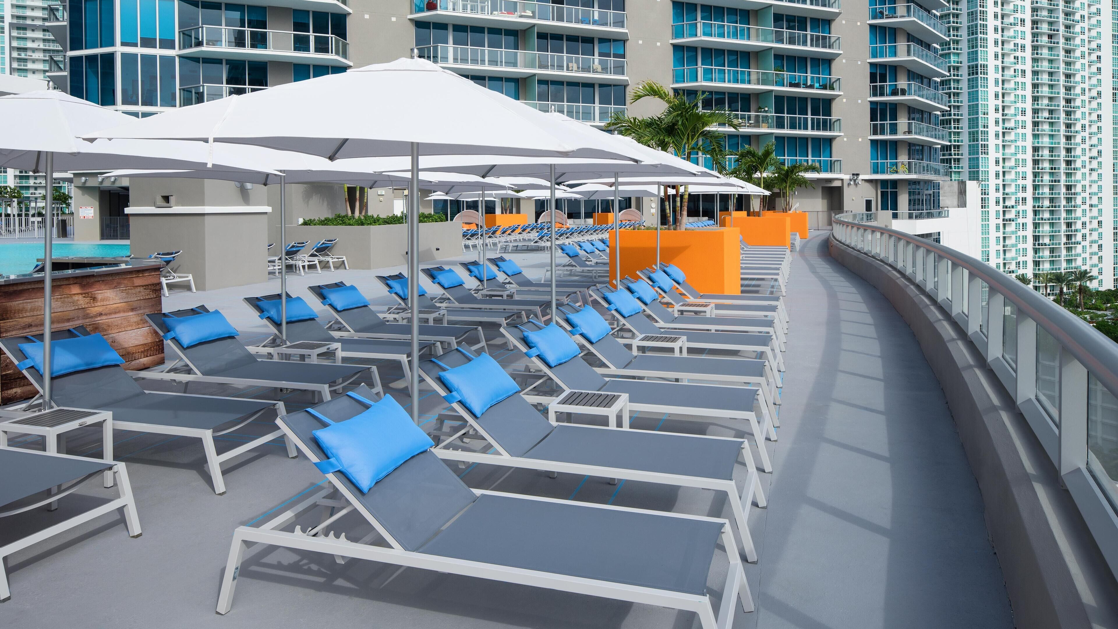 A rooftop lounge area with sunbeds and umbrellas, overlooking a pool and high-rise buildings.