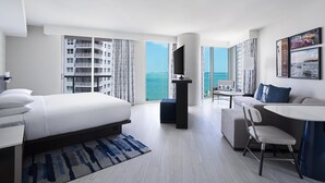 Habitación de hotel moderna con una cama grande, un sofá, una mesita y un televisor. La habitación tiene vista al mar y a un edificio al otro lado del agua.
