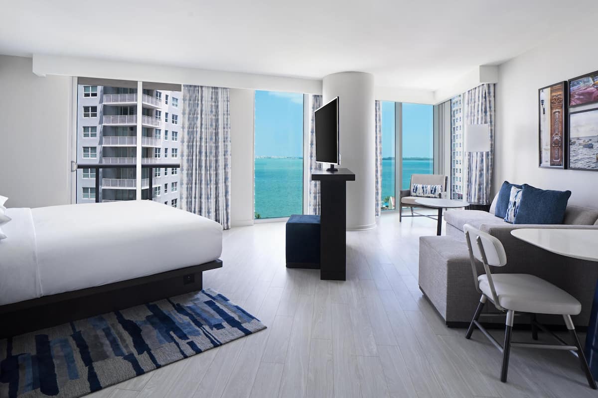 A modern hotel room with a large bed, a sofa, a small table, and a TV. The room has a view of the sea and a building across the water.