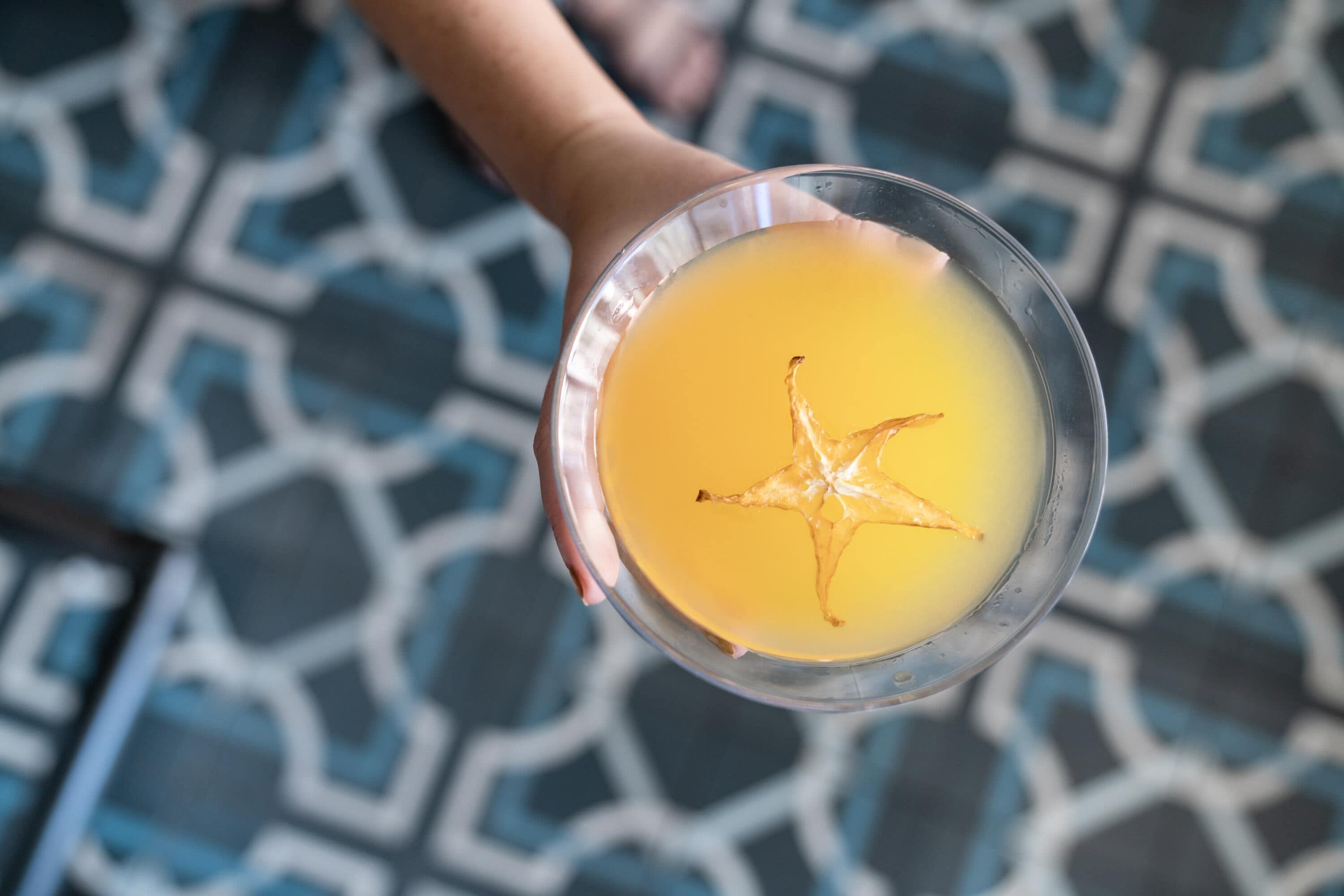 A glass of orange juice with a star-shaped splash on a patterned floor.