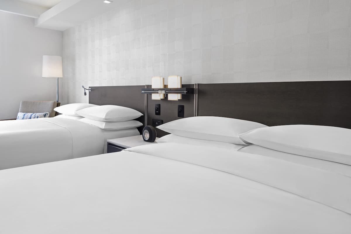 A modern hotel room with two beds, a nightstand, a lamp, and a wall-mounted control panel.
