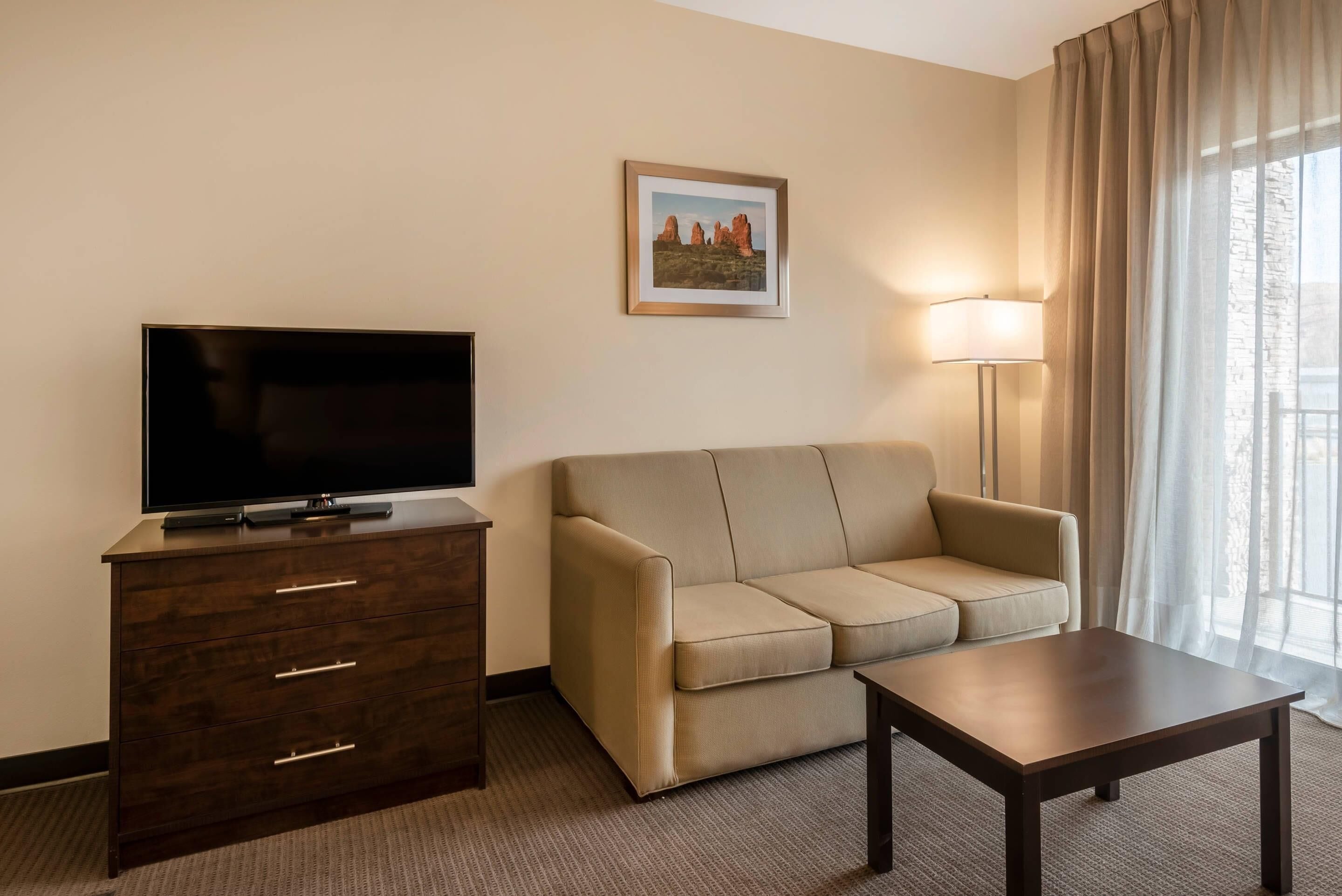 A hotel room with a dark wooden dresser, a beige sofa, a small table, a flat-screen TV, a framed picture on the wall, and a window with curtains.