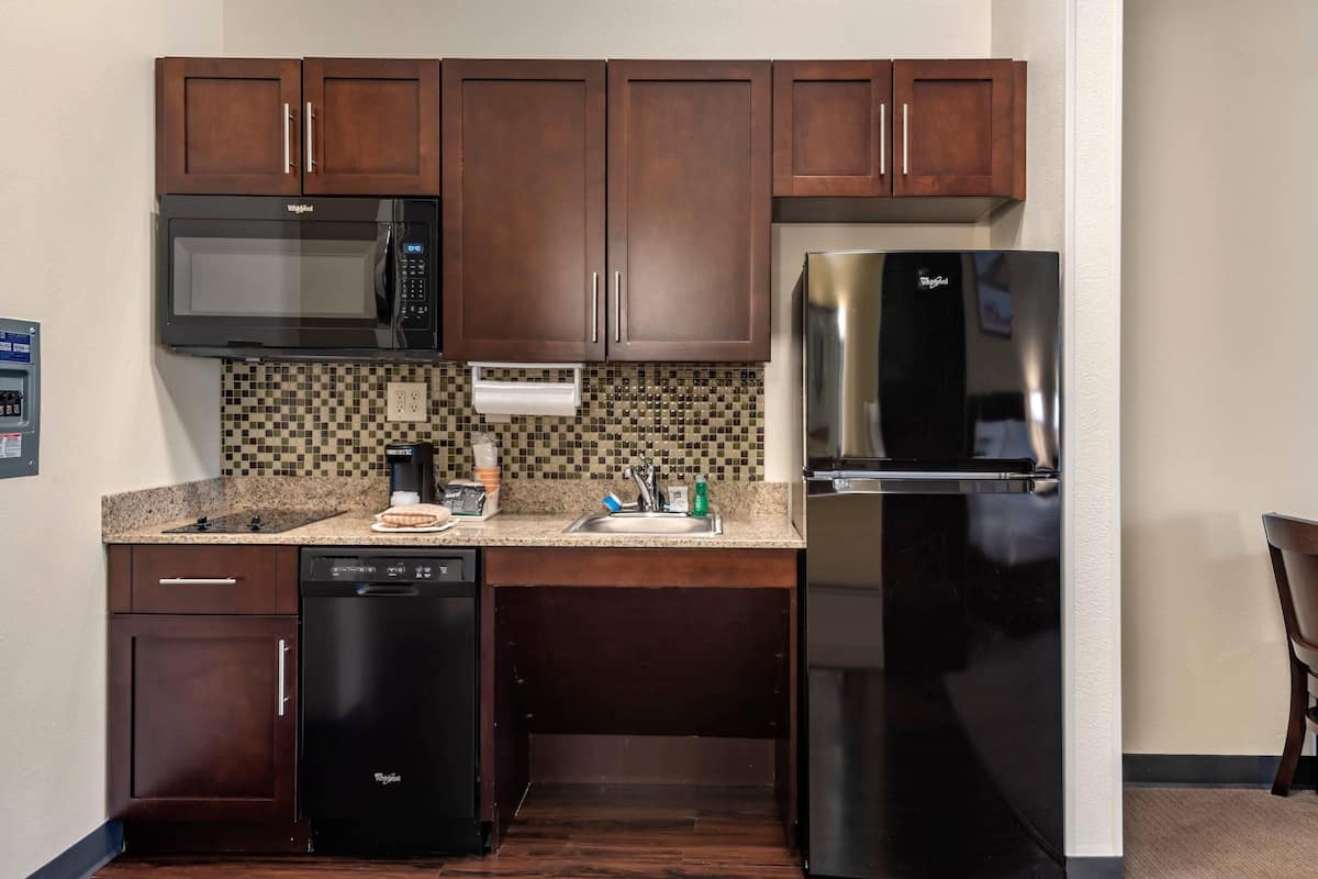 Una cocina compacta con armarios de madera oscura, microondas integrado, refrigerador de acero inoxidable, lavavajillas y salpicadero a cuadros.
