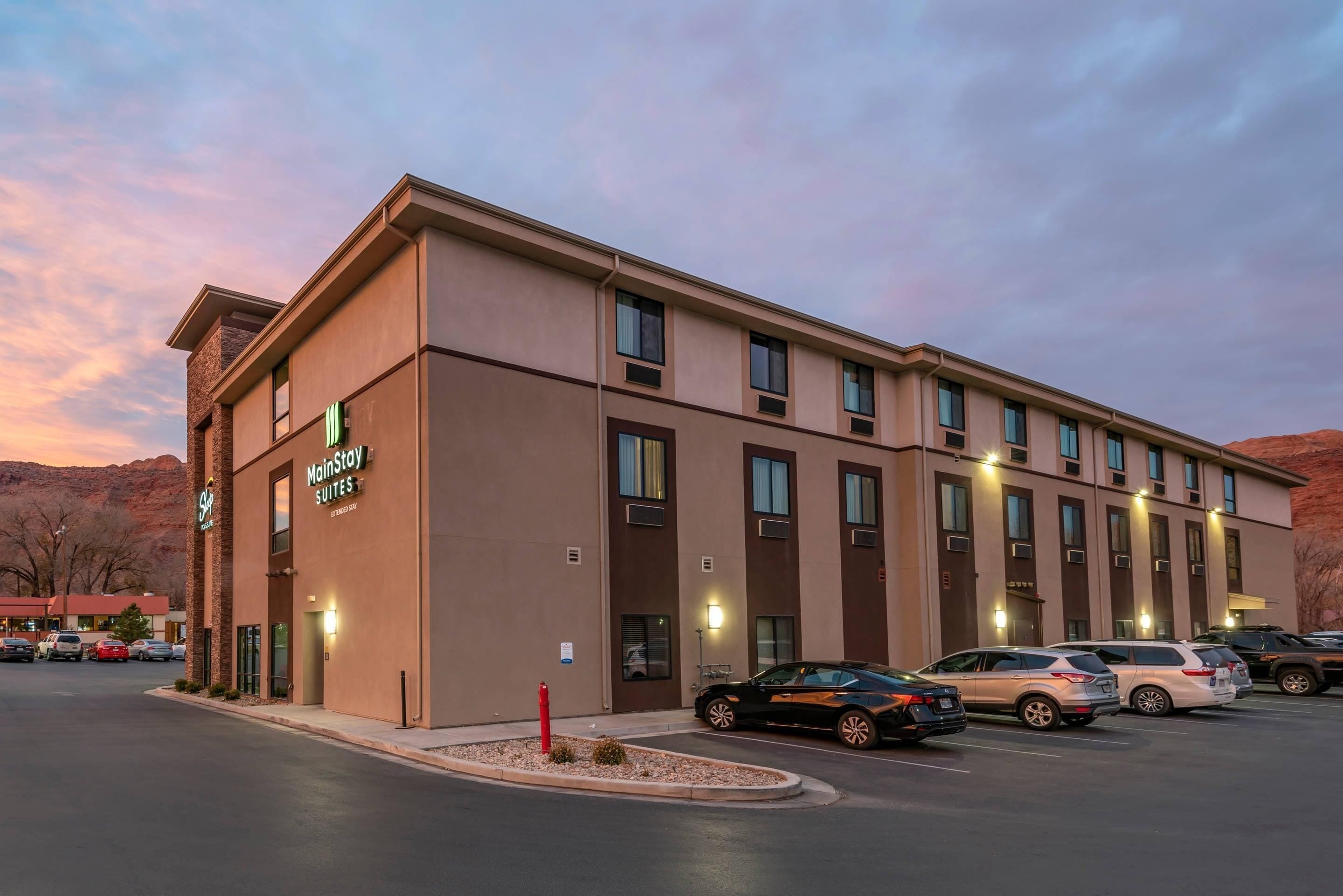A MainStay Suites hotel with a modern design, featuring a parking lot with several cars and a landscaped area with a red fire hydrant.