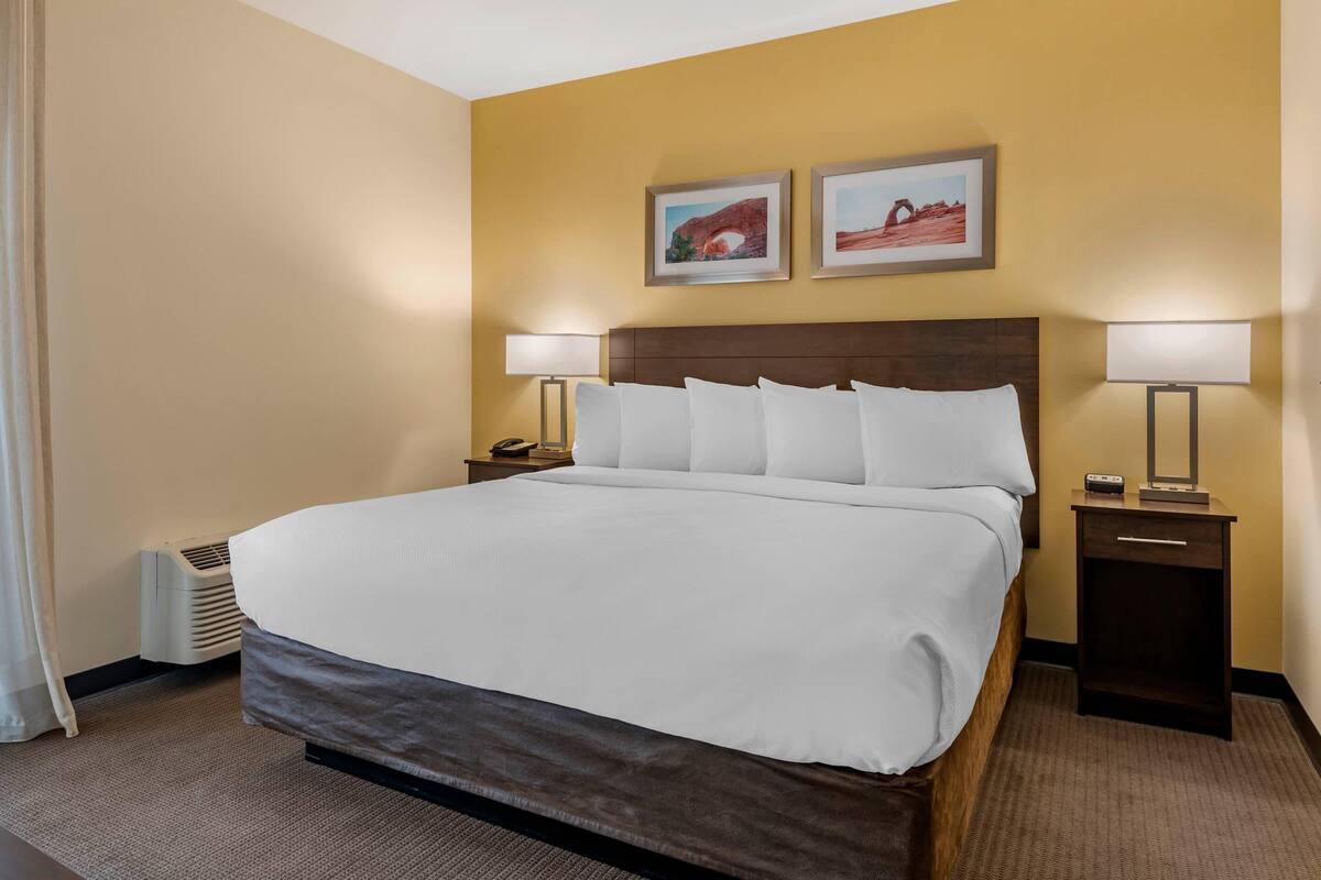 Una habitación de hotel con una cama grande, dos mesitas de noche con lámparas, un aire acondicionado de pared y dos cuadros enmarcados sobre la cama.