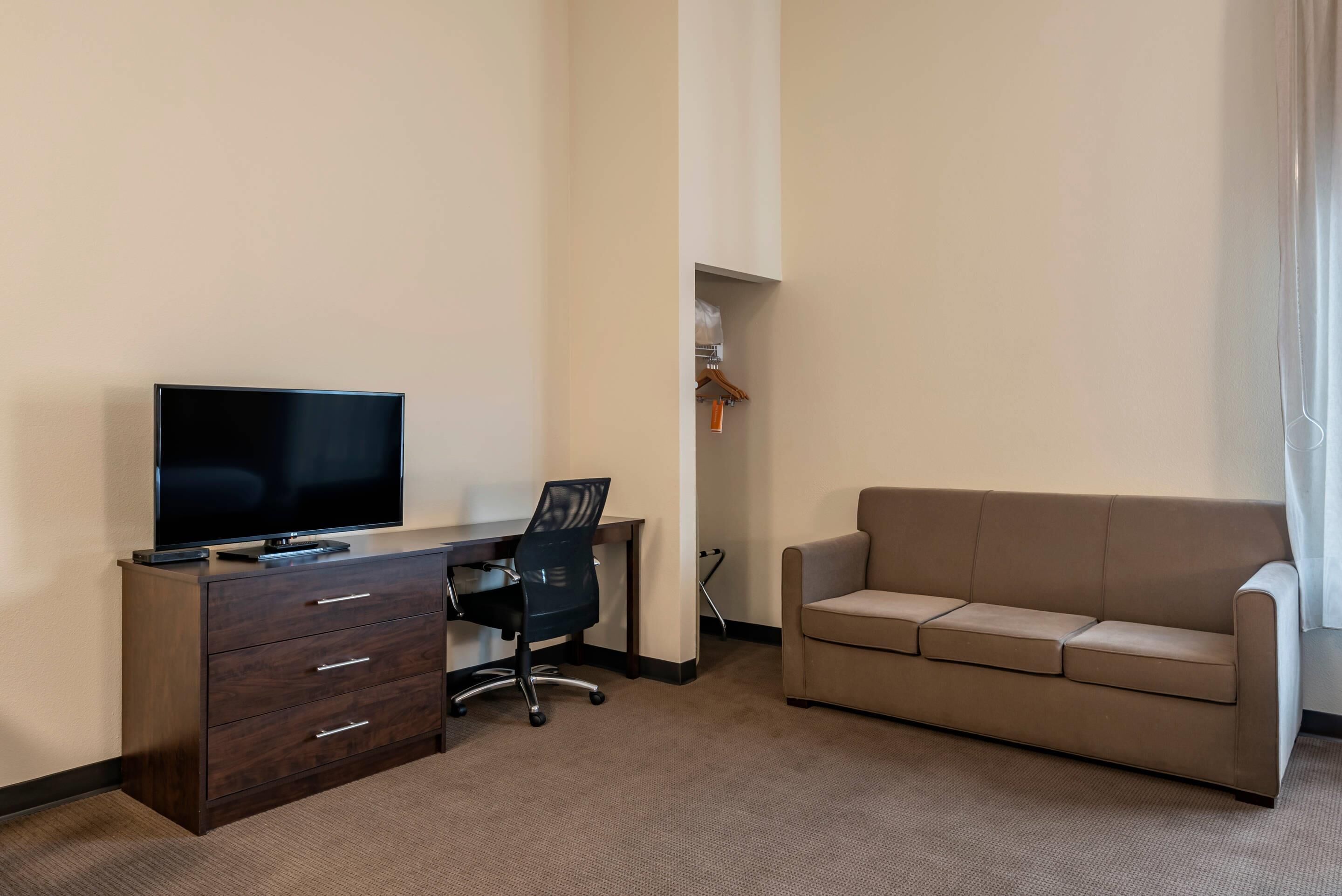 A hotel room with a flat-screen TV on a wooden desk, a black office chair, a beige sofa, and a small closet with a wooden hanger.