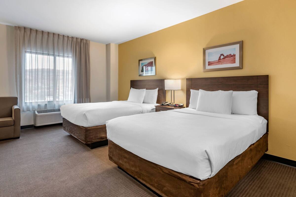 Habitación de hotel con dos camas, una silla, una mesita de noche y un ventanal con cortinas.