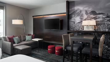 A modern hotel room with a flat-screen TV mounted on the wall, a sofa with red and blue cushions, a small table, and a dining area with chairs.