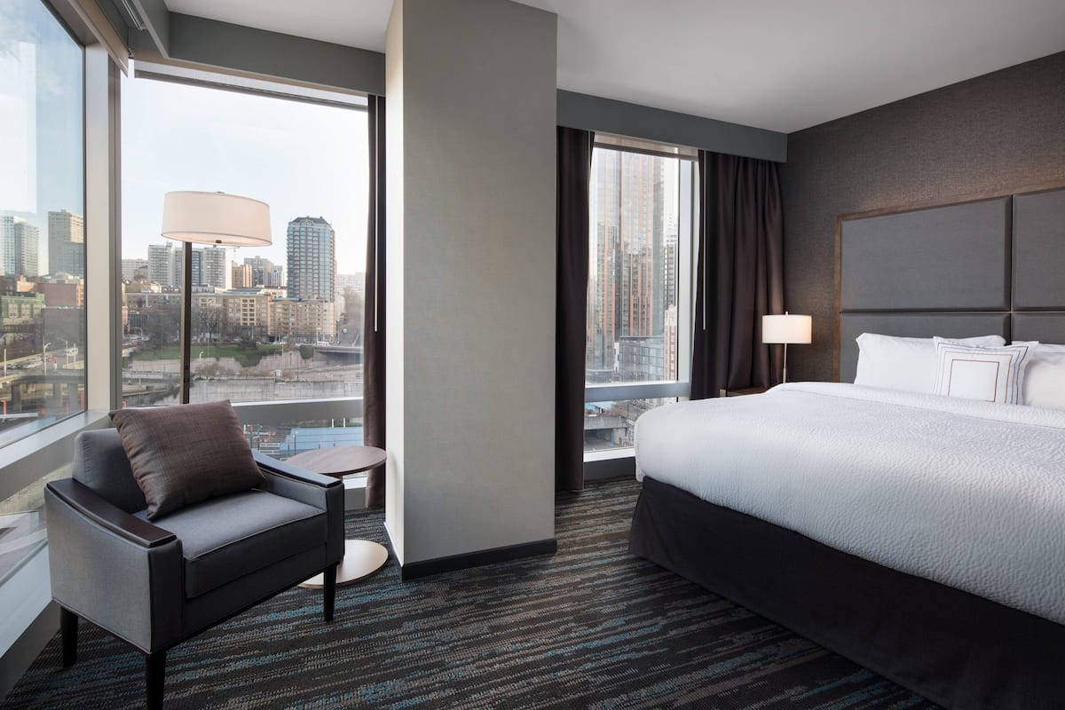A hotel room with a large bed, a chair, a small table, and a view of the city through large windows.