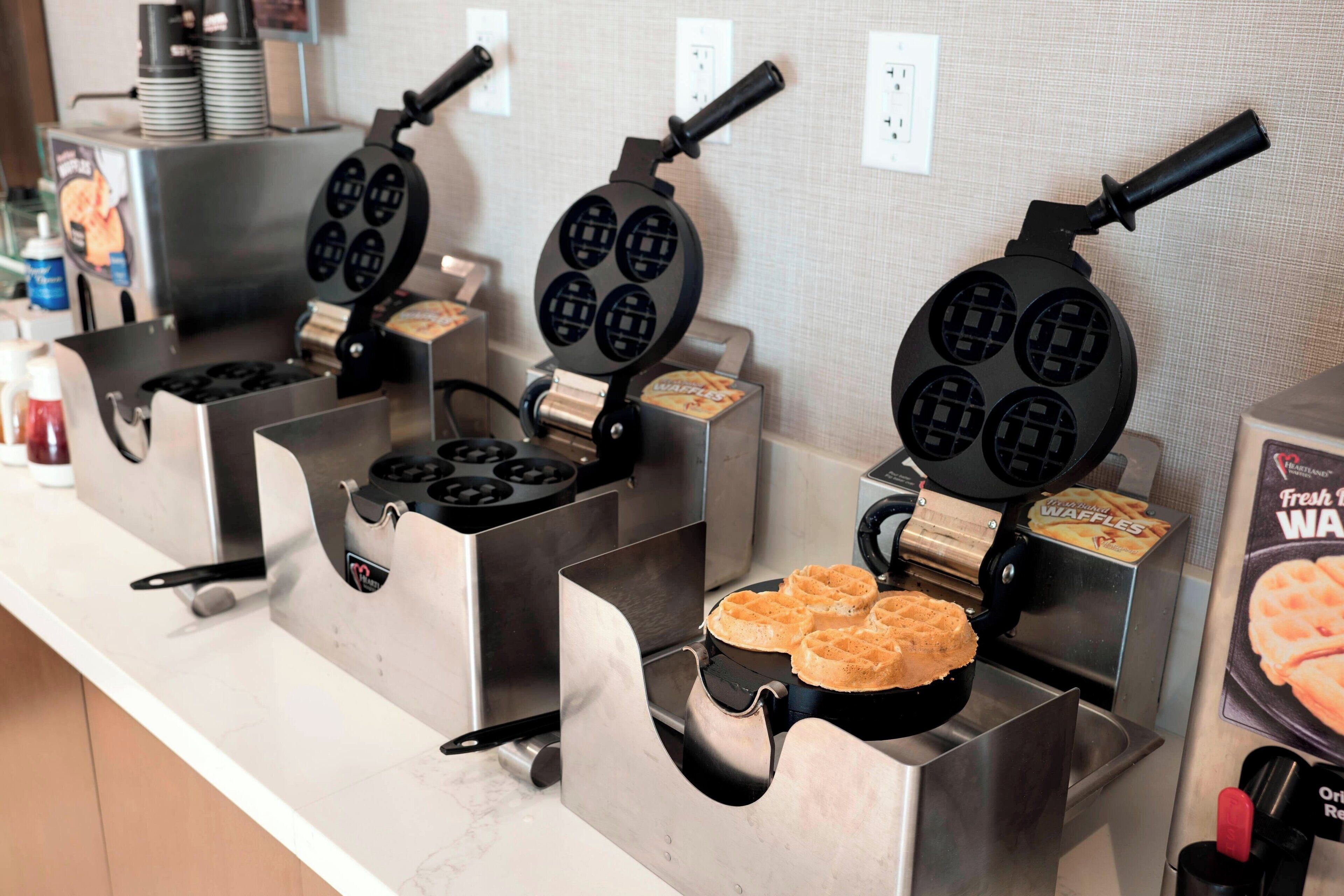 A waffle station with multiple waffle irons in operation, a waffle box, and a bottle of ketchup.