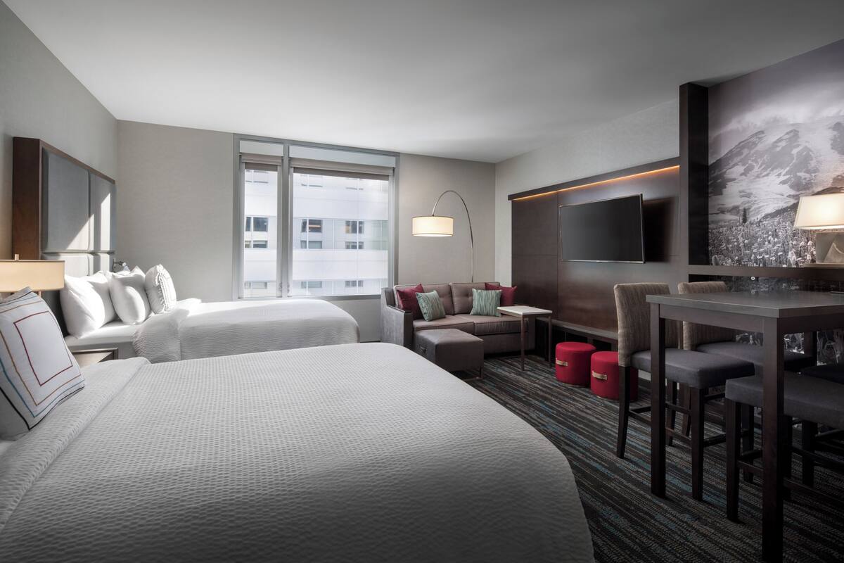 A hotel room with two beds, a sofa, a desk, a TV, and a large window.
