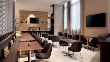 Un restaurante moderno con una barra alargada, varias mesas y sillas. Hay un televisor de pantalla plana montado en la pared e iluminación colgante sobre la barra.