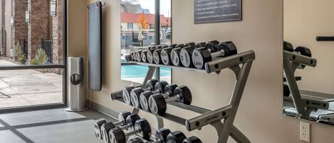 A fitness center with dumbbells on racks, a safety rules sign, and a view of a building and trees outside.