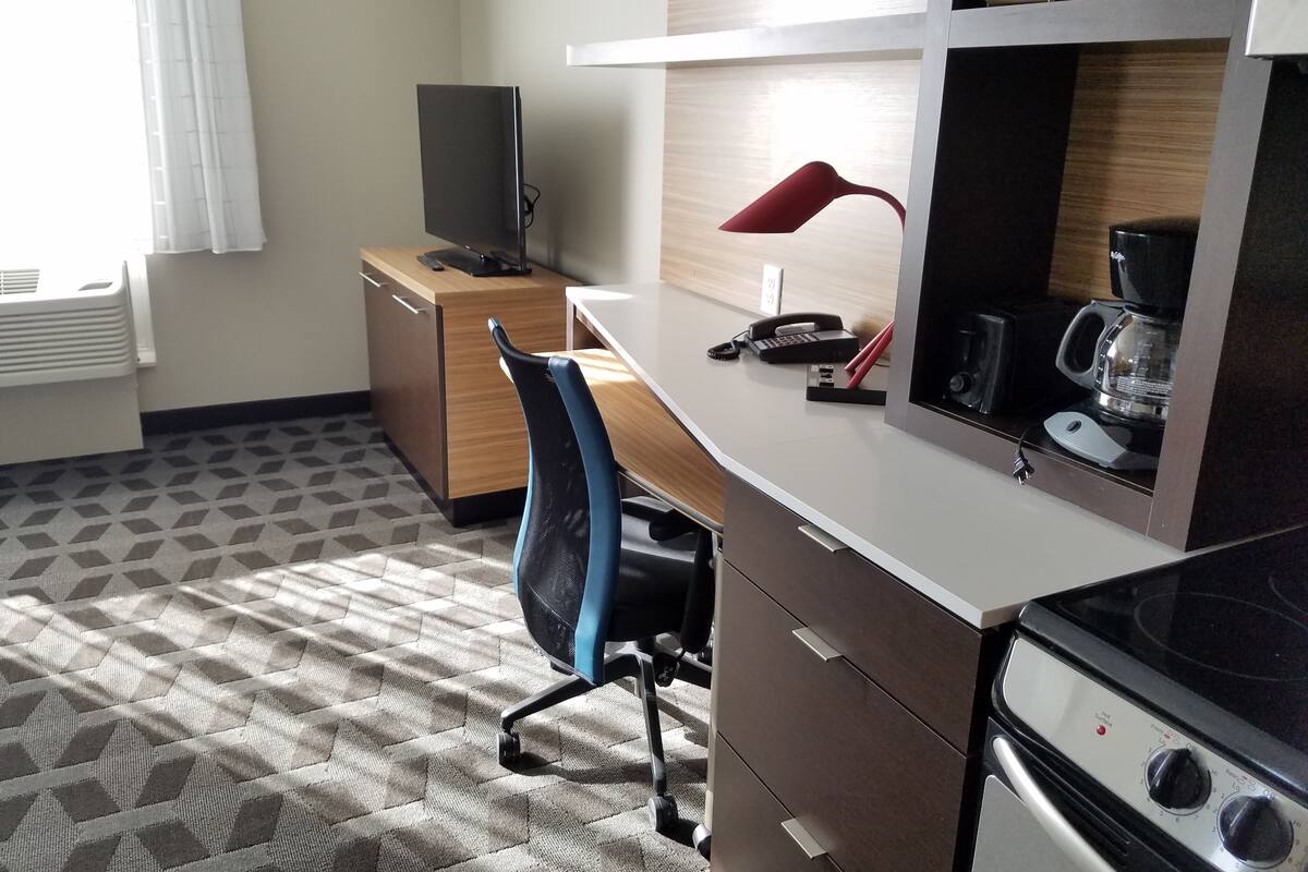 A hotel room with a kitchenette, a desk, a chair, a TV, a red lamp, and a coffee maker.