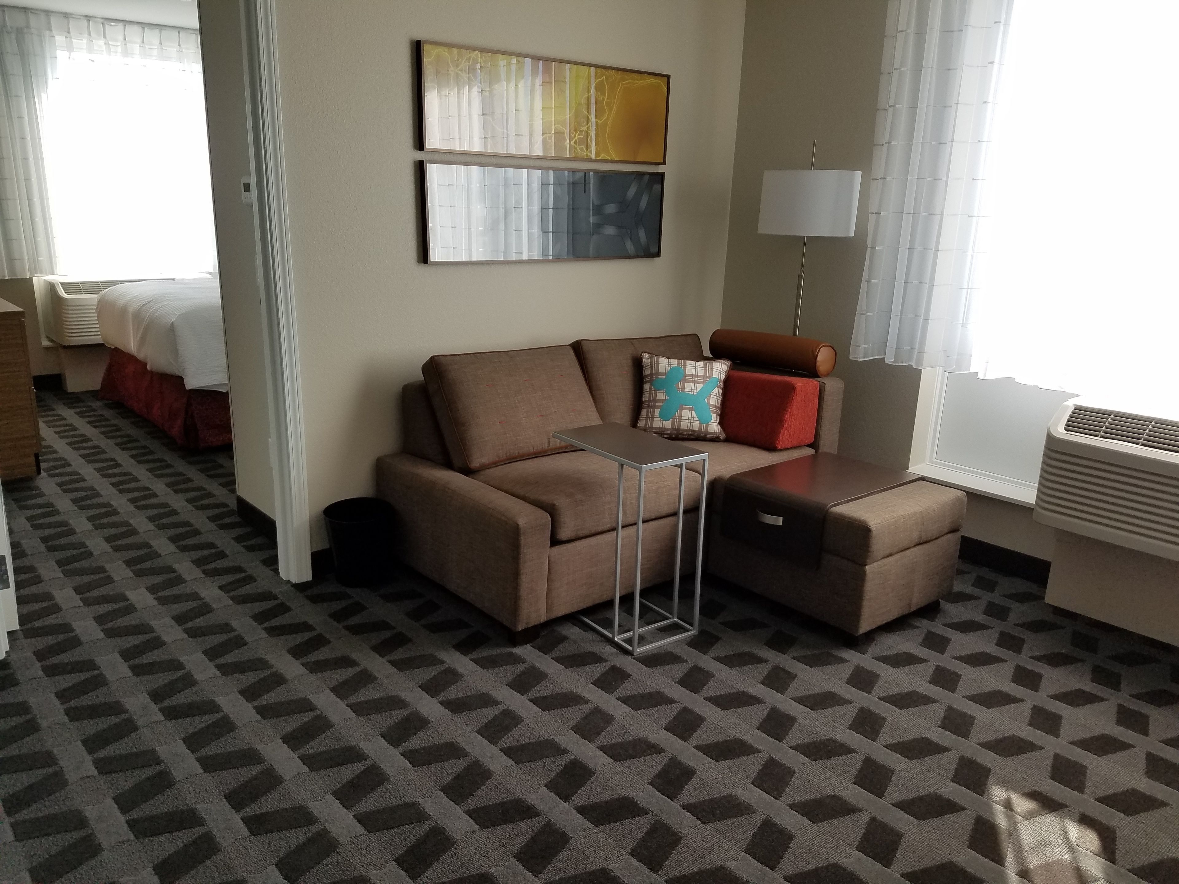 Quarto de hotel com sofá marrom, mesinha de apoio, cama com colcha vermelha e janela com ar condicionado branco.