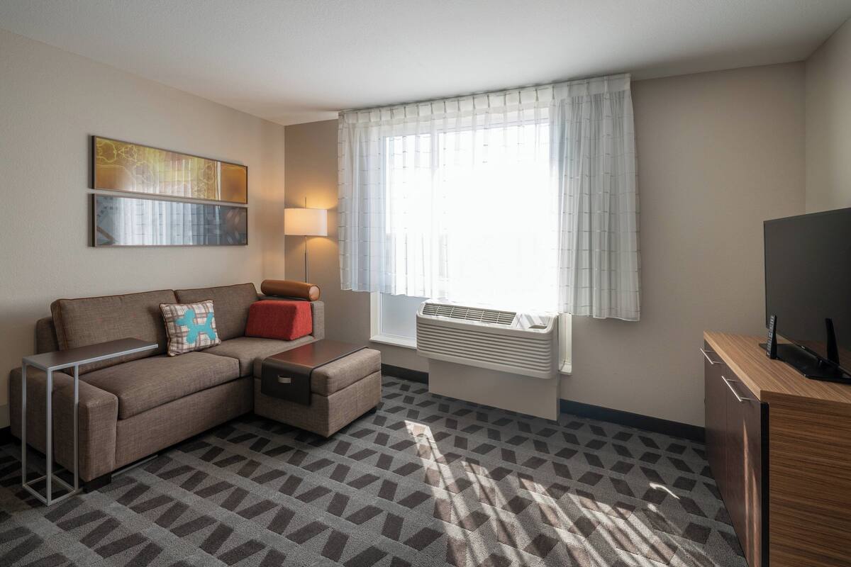 A modern hotel room with a sofa, a small table, a lamp, a TV, and a window with curtains.