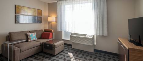A modern hotel room with a sofa, a small table, a lamp, a TV, and a window with curtains.
