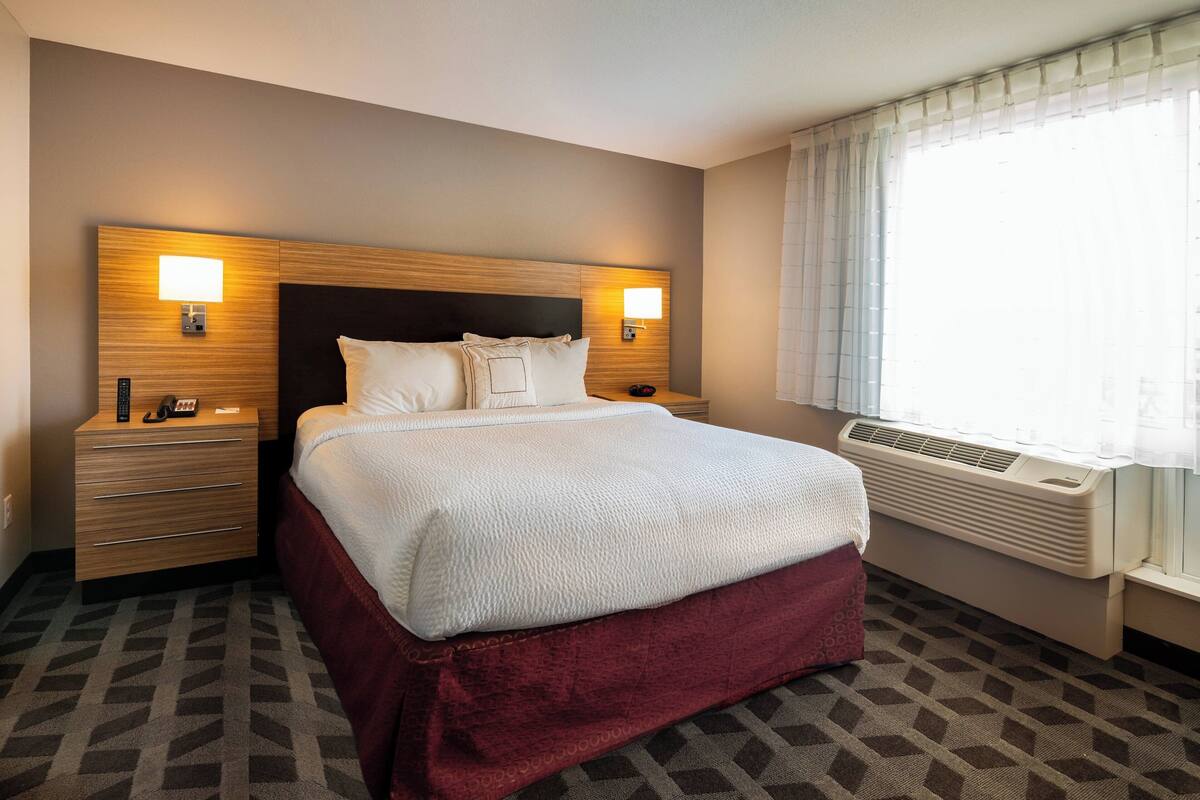 A hotel room with a large bed, two bedside tables with lamps, a window with curtains, and a wall-mounted air conditioner.