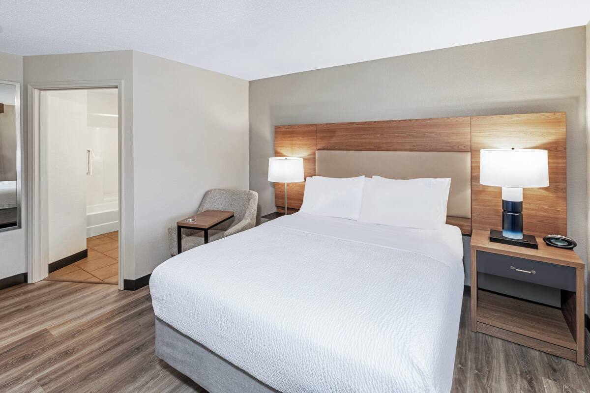 Une chambre d’hôtel avec un grand lit, deux tables de chevet avec des lampes, une chaise et une tête de lit en bois.