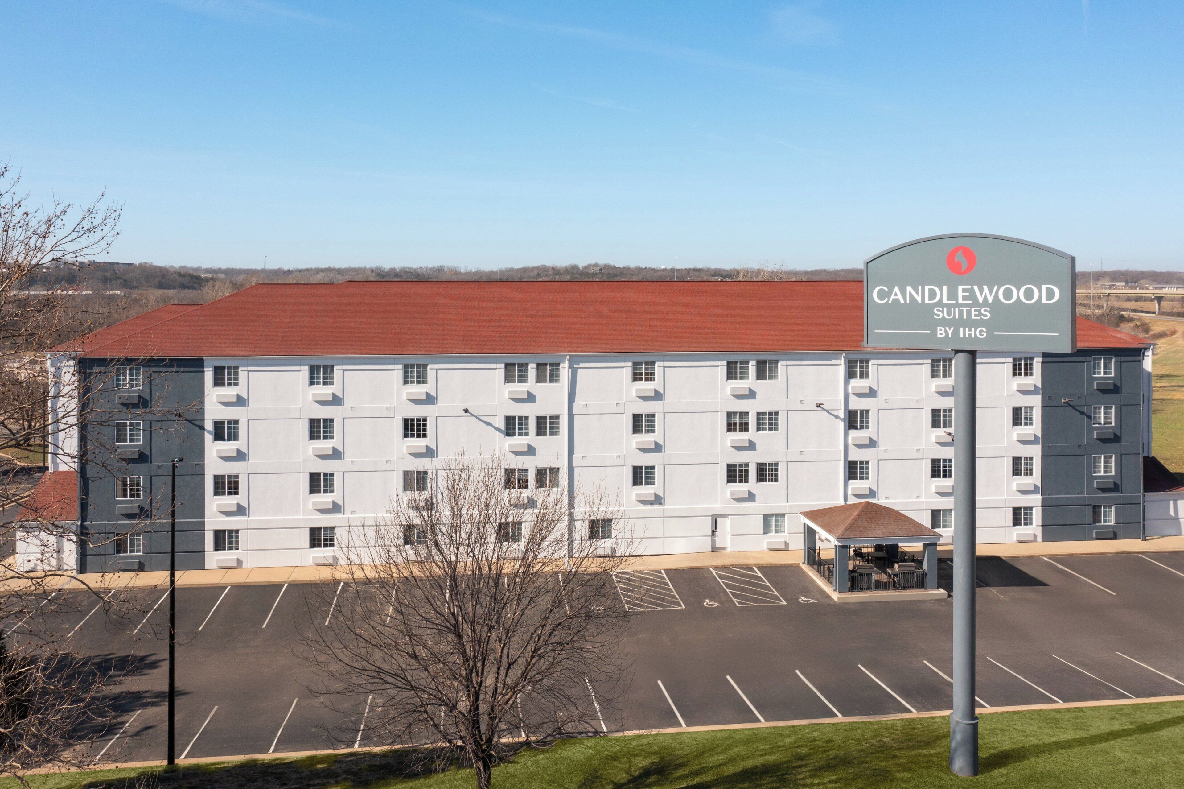 A hotel with a large parking lot, a covered outdoor seating area, and a sign indicating it is Candlewood Suites by IHG.