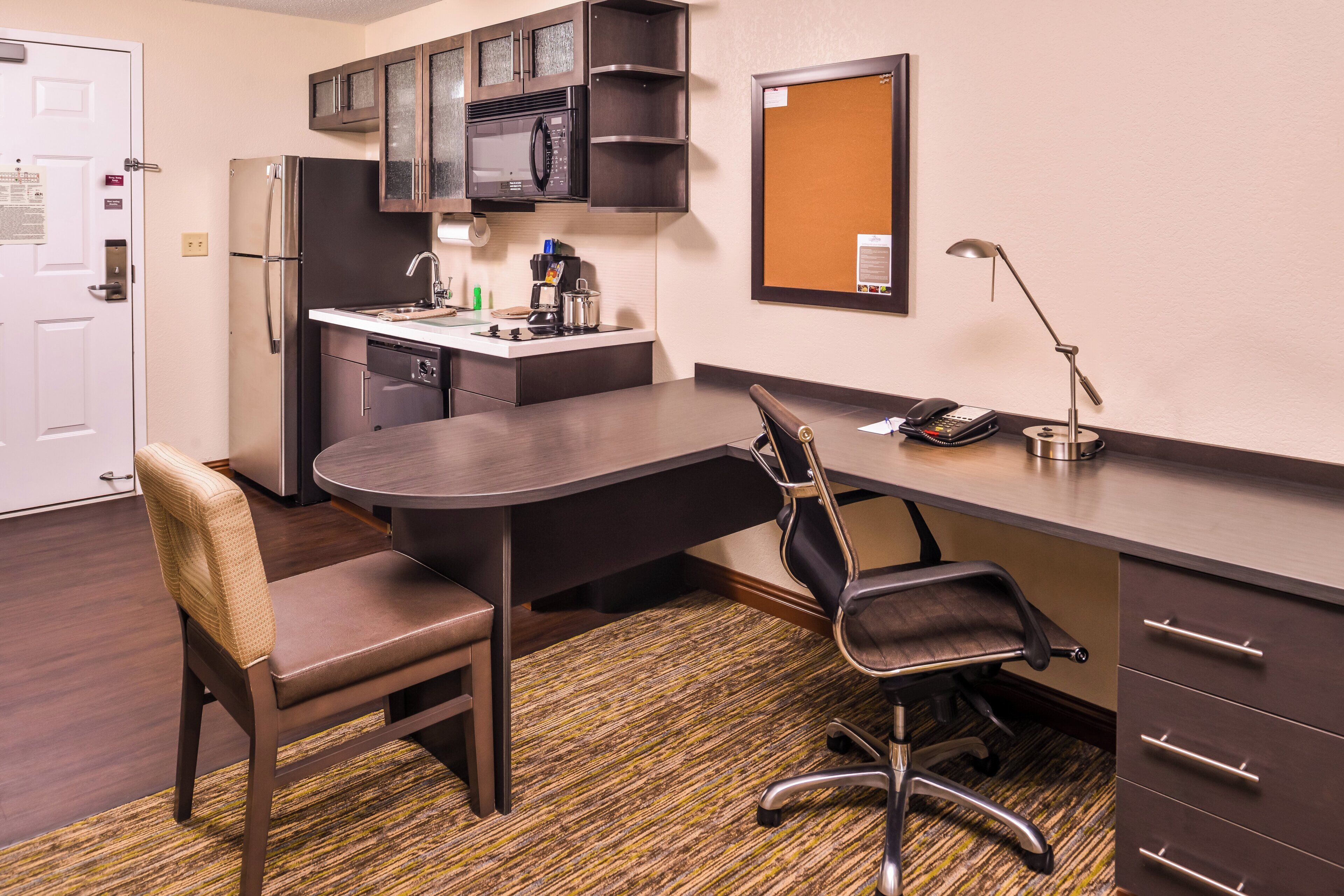 A compact hotel room with a kitchenette, a dining area with a table and chairs, a desk with a chair, a telephone, and a bulletin board.