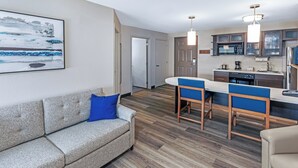 A modern kitchen with a microwave, stove, and dishwasher. There is a dining area with a bar and two blue chairs. A gray sofa with a blue pillow is in the living area.