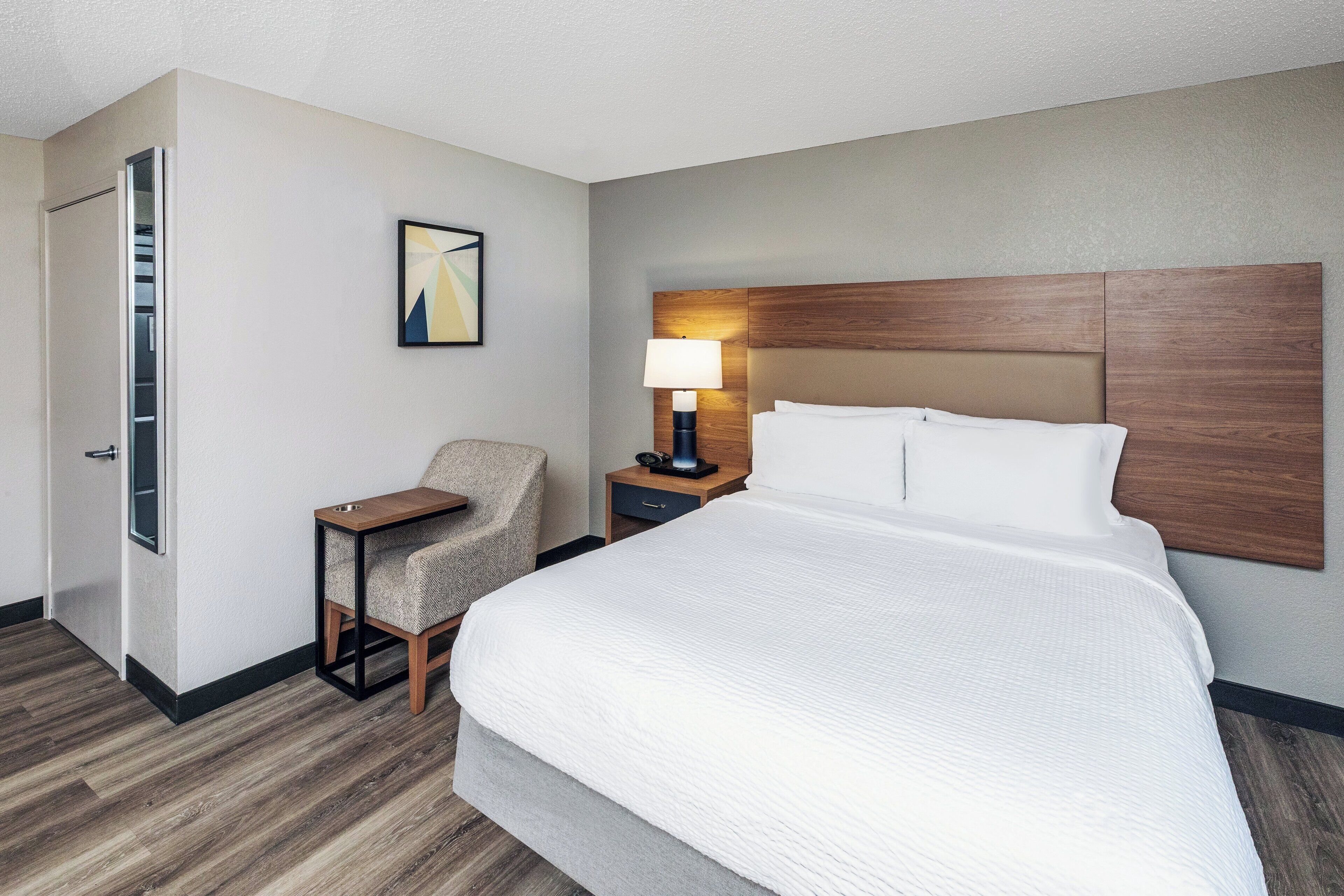 A hotel room with a large bed, a chair, a nightstand with a lamp, and a wall-mounted shelf.