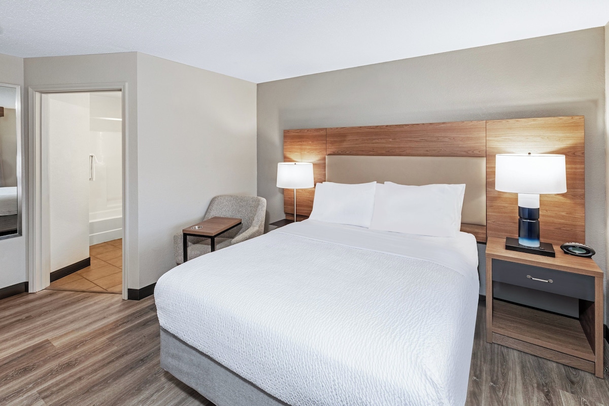 Une chambre d’hôtel avec un grand lit, deux tables de chevet avec des lampes, une chaise et une tête de lit en bois.