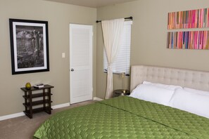 A bedroom with a green quilted bedspread, a framed black and white photo, a white door, a bedside table with a lamp, and a colorful striped artwork on the wall. - Encanto Vistas, A Golf Property in Central Phoenix (Phoenix)