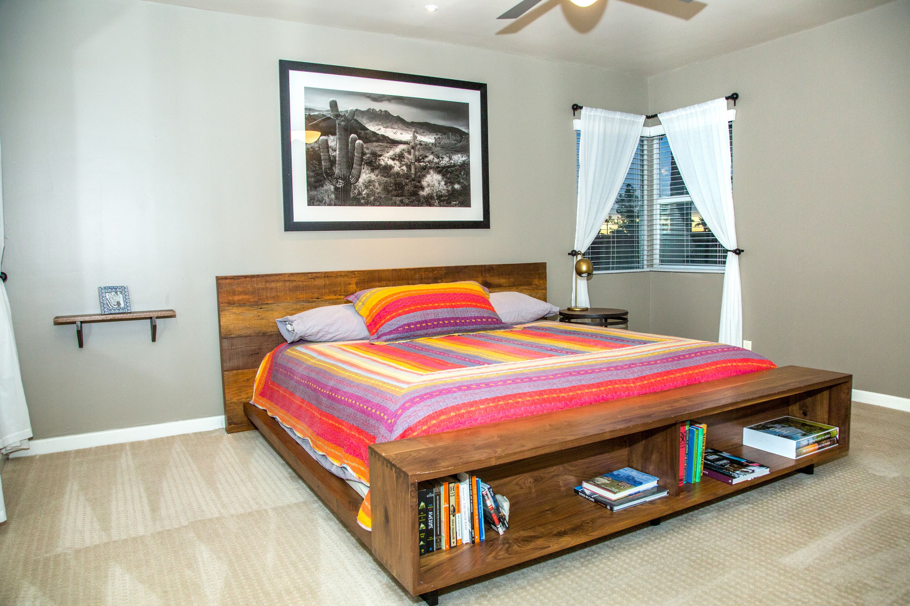 A bedroom with a wooden bed, a colorful blanket, a bookshelf, a framed picture on the wall, and a window with curtains.