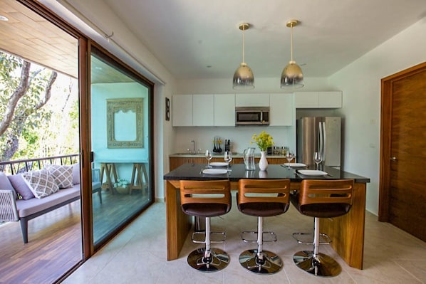 A modern kitchen with a central island, bar stools, pendant lighting, and a view of the outdoors.