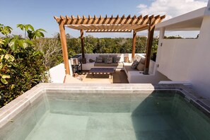 Ett poolområde med en pergola av trä, sittplatser och utsikt över grönskan.