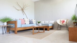A modern living room with a light blue sofa, a wooden coffee table, and a wicker chair. There are potted plants and a wall-mounted decor piece. - Alquimia 2 bedroom apartment sleeps 4 (Tulum)
