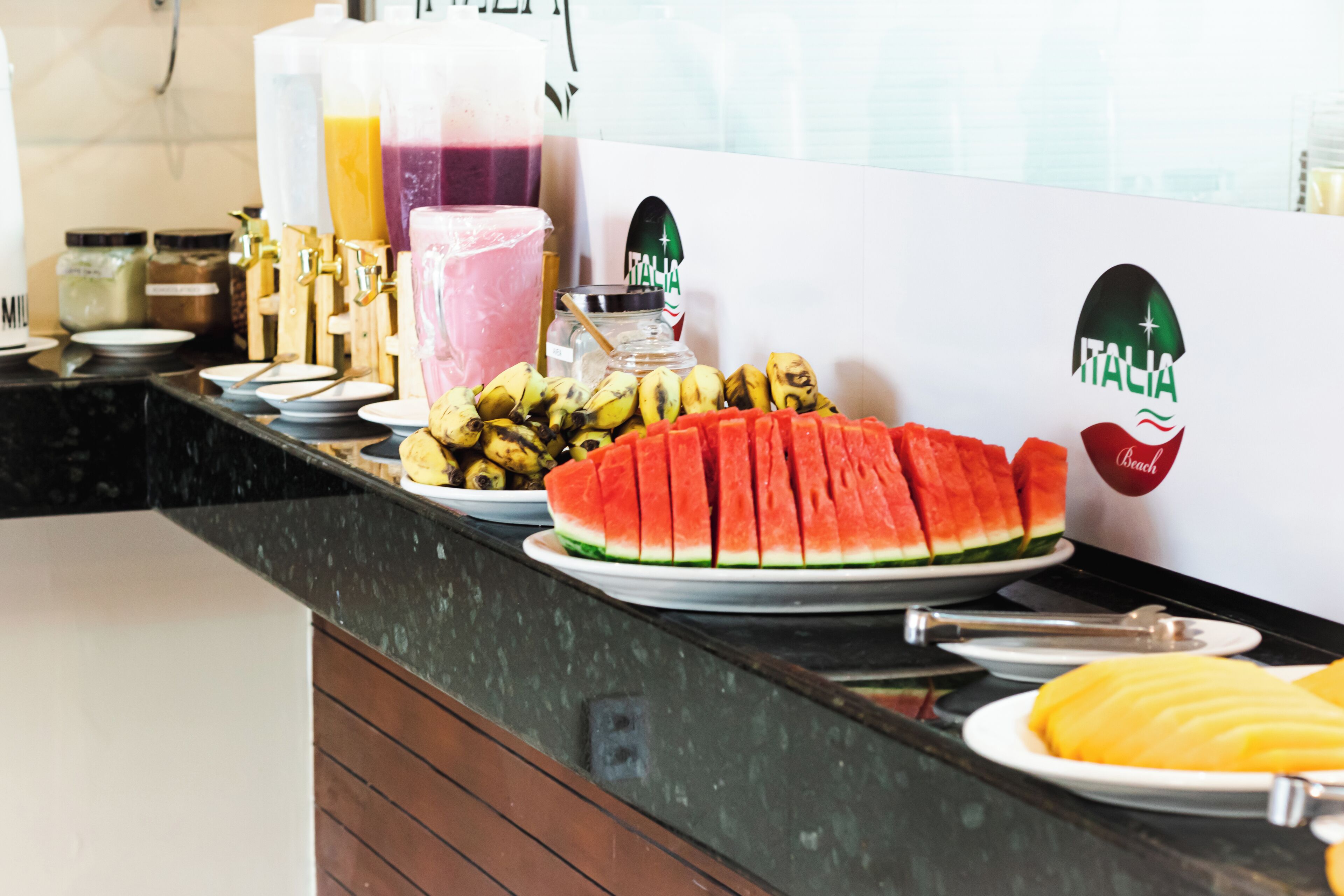 A breakfast buffet with various fruits, a watermelon, and a juice dispenser.