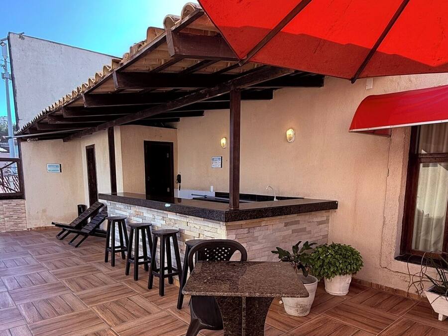 Un patio exterior con piso de madera, una zona de bar con taburetes y una sombrilla roja que da sombra.
