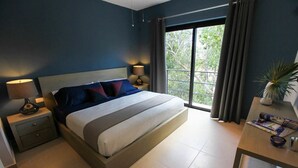 A modern bedroom with a large bed, two bedside tables with lamps, a view of greenery through a large window, and a small table with a plant. - Tony Condo 2 bedroom sleeps 4 (Tulum)