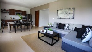 A modern living room with a grey sofa, black and white cushions, a coffee table, and a dining area with a white table and chairs. - Martiki Condo 2 bedroom sleeps 4 (Tulum)
