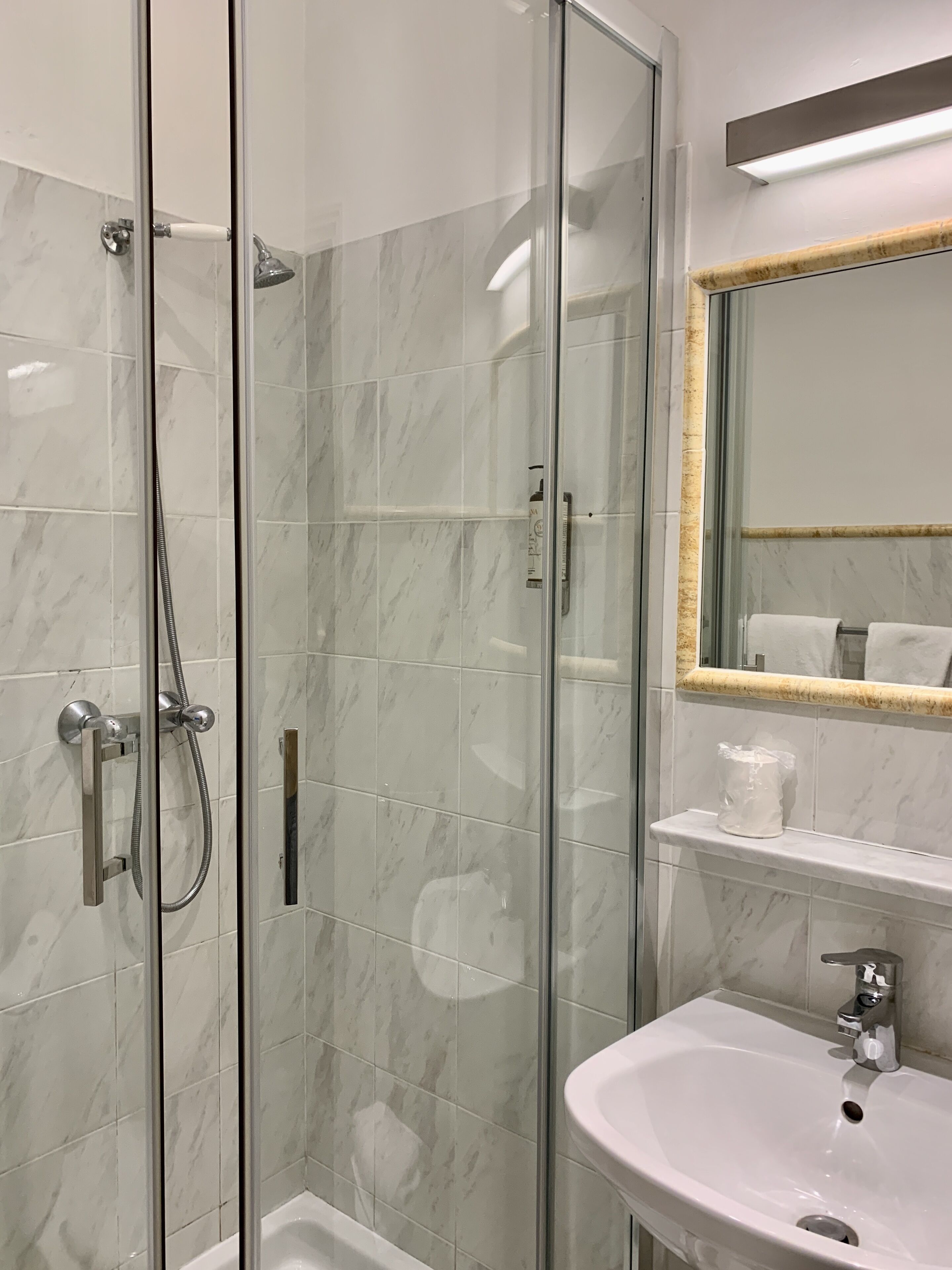 Ein modernes Badezimmer mit einer gläsernen Duschkabine, einem weißen Waschbecken und einem Spiegel mit goldener Rahmung.