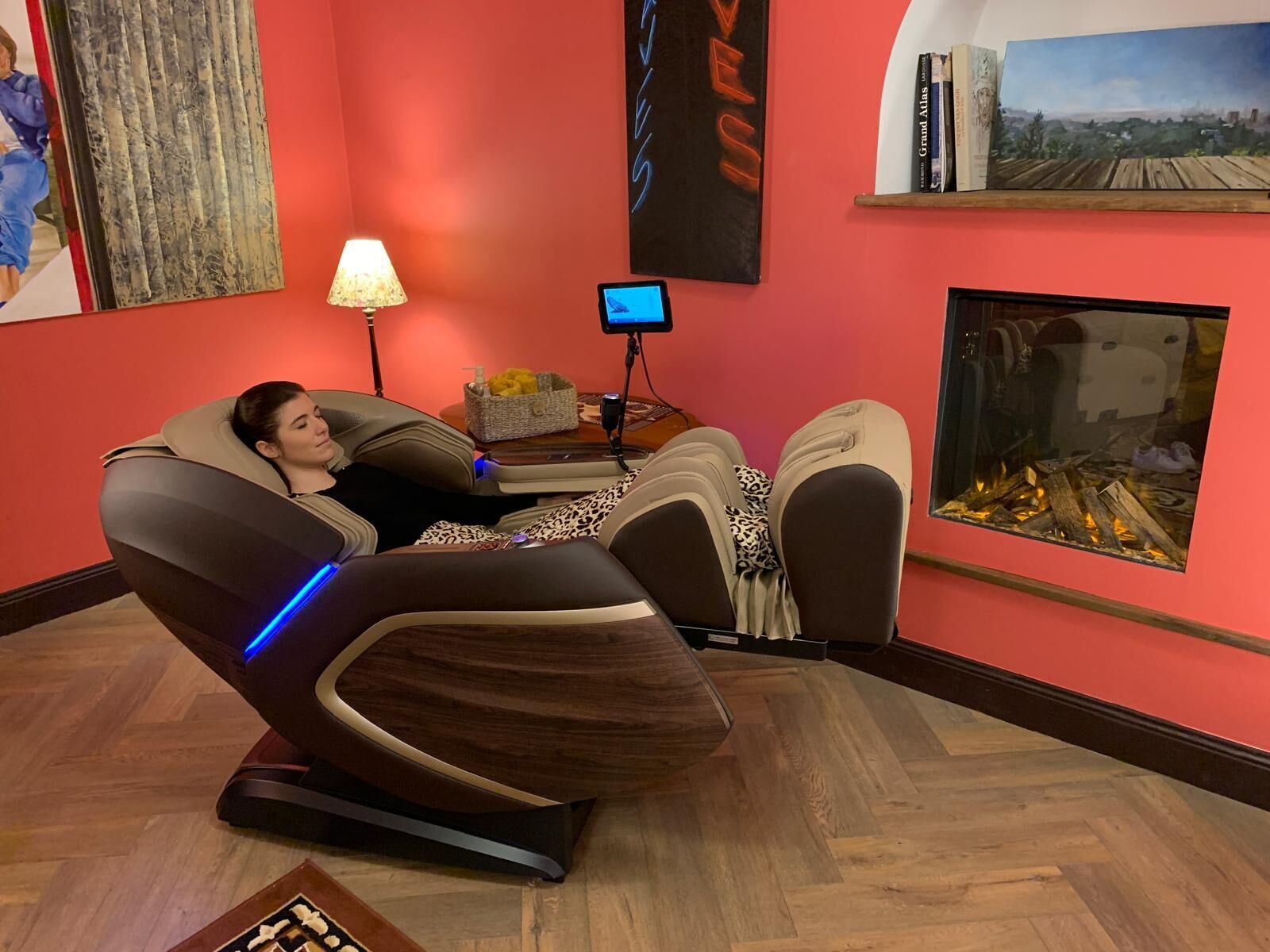 En massagestol i et rum med rød væg, en lampe, et lille bord med en kurv og en pejs.