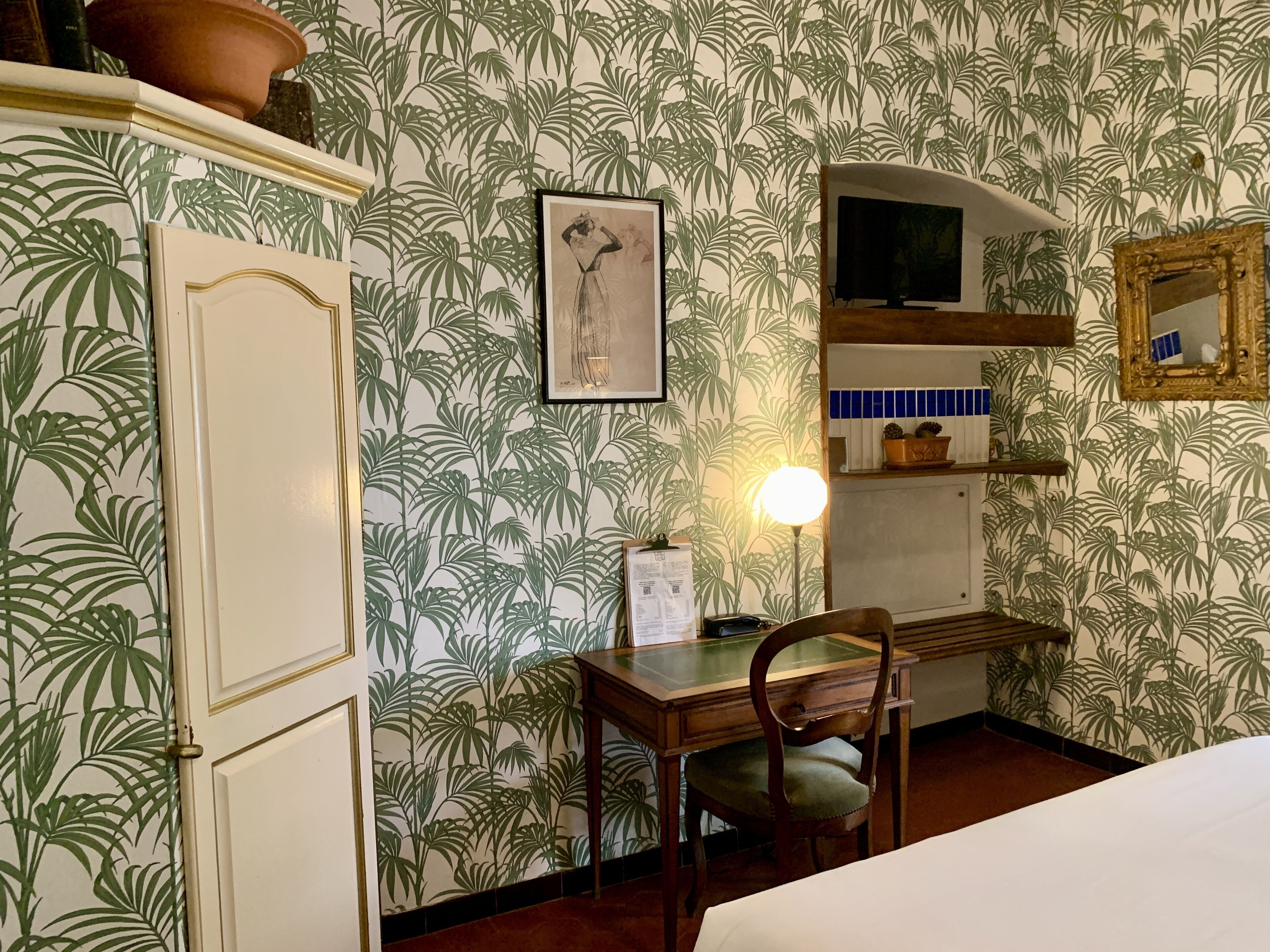 Um quarto com papel de parede estampado, uma escrivaninha com cadeira, uma cama, uma televisão e um espelho na parede.