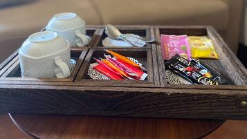 A wooden tray with white cups and colorful snacks on a table in a living room with a beige sofa and a red blanket in the background.