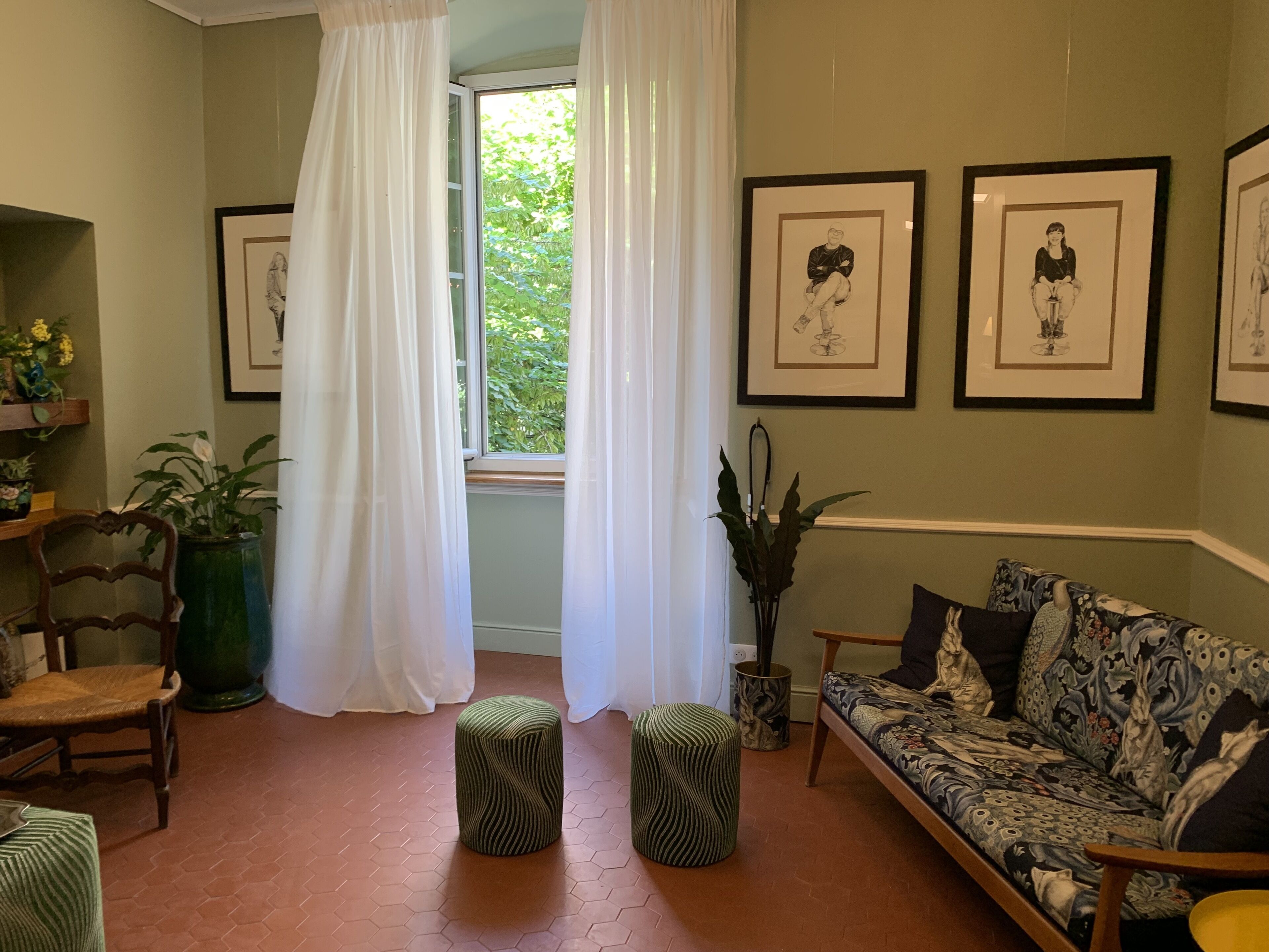Un salon comprenant un canapé, deux poufs, un fauteuil, une fenêtre avec des rideaux et des tableaux encadrés accrochés au mur.