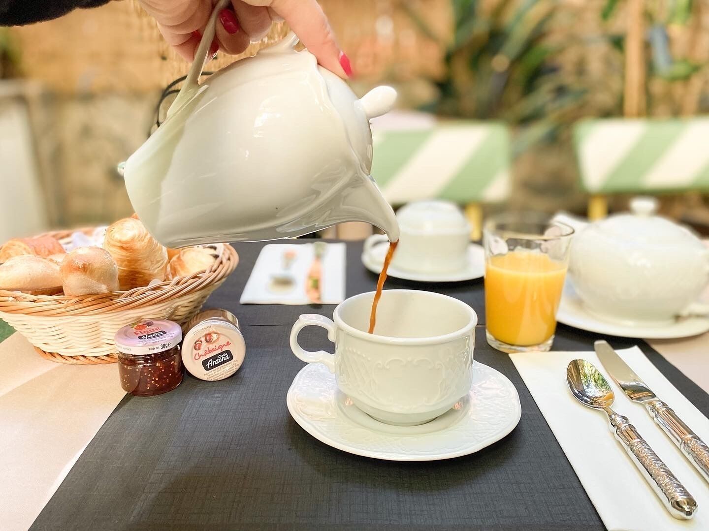 A breakfast setup with a teapot pouring coffee into a cup, a basket of bread, a glass of orange juice, and a plate of pastries.