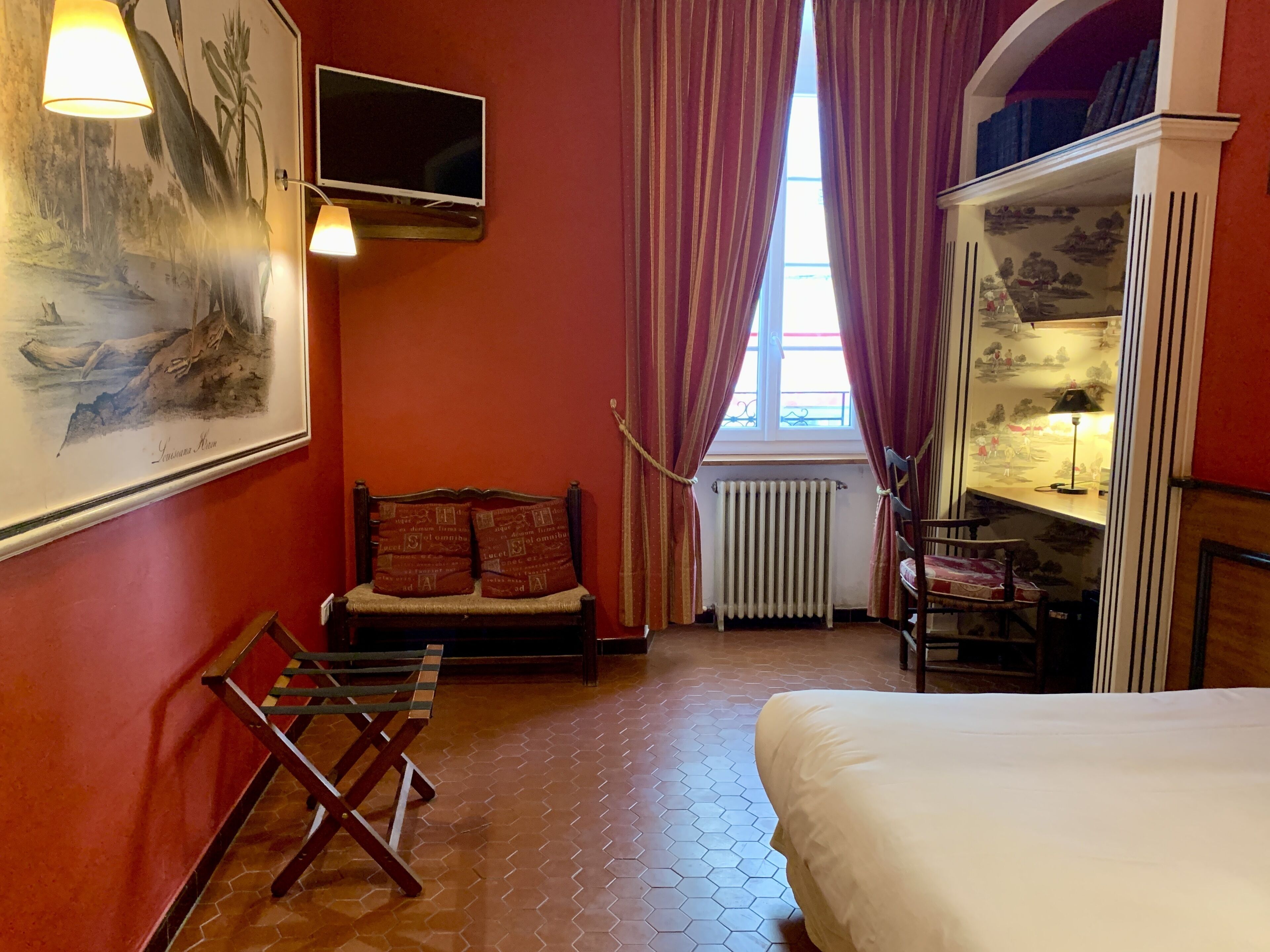 Ein Zimmer mit einem Bett, einem Sessel, einem Schreibtisch mit Lampe, einer Heizung, einem Fenster mit Vorhängen und einem Bild an der Wand.