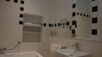 Une salle de bain avec une baignoire, un lavabo et des toilettes. Il y a des étagères au-dessus de la baignoire et une poubelle près des toilettes.